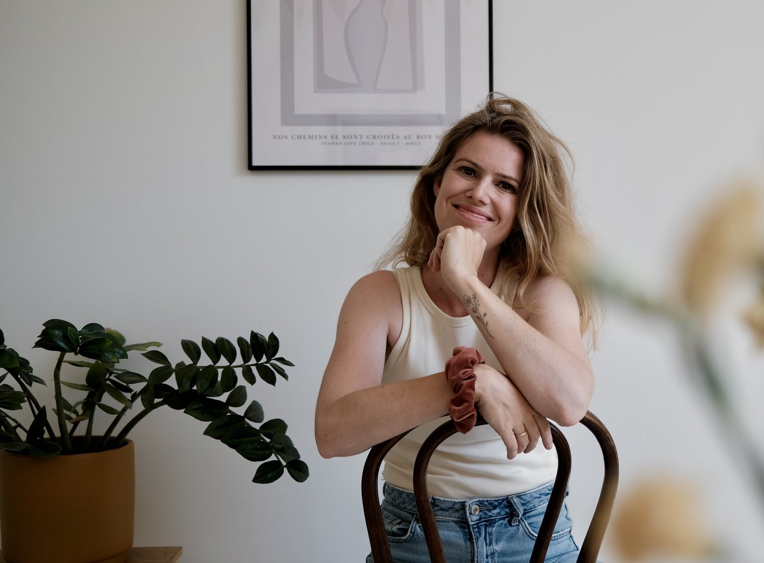 Anje van Gelder op een houten stoel in haar atelier, met een kunstposter aan de muur